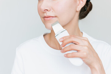 Young Beautiful Caucasian brunette woman using a white ultrasonic facial cleaning device. Cosmetic...