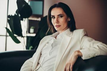 Professional businesswoman in a white suit sits confidently in a modern office with natural...