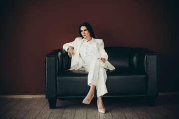 Stunning businesswoman sitting on a modern black sofa, dressed in elegant white formalwear, against...