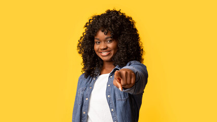 Cheerful young black woman pointing at camera on yellow studio background, panorama with copy space. Positive african american pretty lady flirting, smiling and pointing at camera