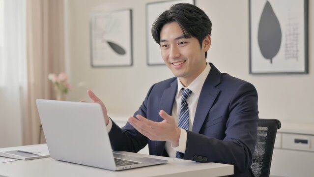 Happy Asian businessman in suit talking on laptop, representing professionalism, communication and technology integration.
