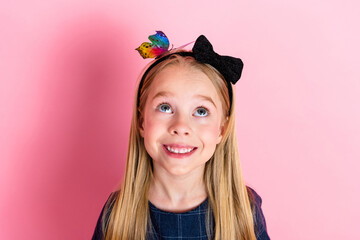 Charming young girl with a black bow headband and colorful butterfly on pink background, looking up...