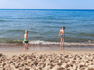 Two young boys stand quietly at the edge of the sea, watching gentle waves touch their feet — a...