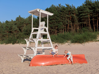 Two young boys play near a white lifeguard tower and overturned orange rescue boat on a quiet sandy...
