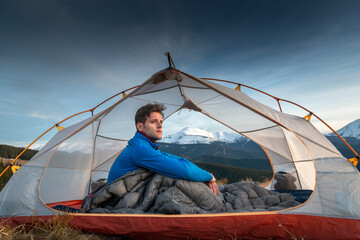 Male camper sitting in sleeping bag inside tent, looking at snowy mountain landscape at sunrise
