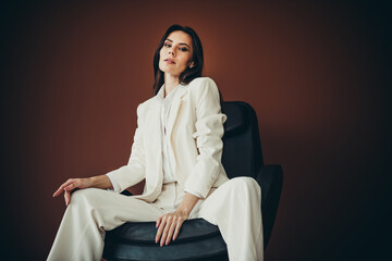 Stunning businesswoman in white suit sitting stylishly on chair against a brown background exuding...