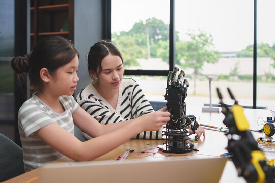 Two Diverse teenage girls wearing striped sweater assembling and programming a robotic arm in a modern classroom with focus and teamwork, surrounded by electronic components.