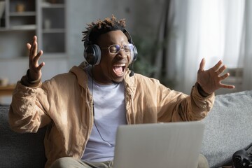 Ecstatic young man wearing a headset shouts and gestures in celebration of a gaming win An African American individual enjoys using a laptop and joystick in their free time