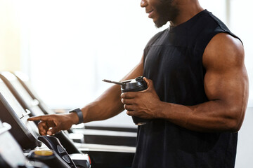 Cropped of muscular african american man holding sports drink, standing on treadmill, drinking...