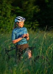 Young cute woman cyclist in uniform, helmet stands with gravel bike among tall grass in rays of...