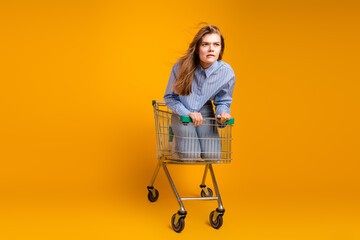 Young woman sitting in a shopping cart against yellow background expressing uniqueness and...
