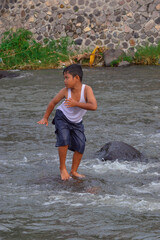 A young boy strikes a pose while standing on a large rock partially submerged in the flowing river,...