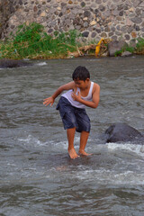 A playful moment of a boy performing a balancing act on a slippery river rock, arms extended to...