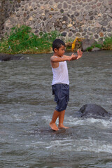A boy maintains his stance atop a slippery river rock, arms raised for balance while standing...