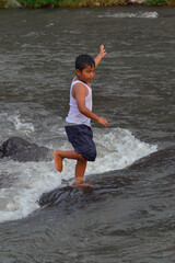 A young boy stands confidently with one foot raised on a stone in the river, performing a playful...
