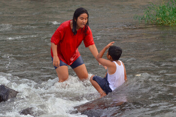 While standing in the flowing river, a teen girl speaks to a boy seated on a stone, his head in his...