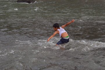 Fototapeta premium A young boy leans forward with energy, joyfully splashing water with both hands in the middle of the rapidly flowing river current.