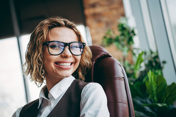 Charming businesswoman in formal attire smiling in a modern office space with plants and stylish...