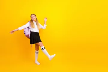 Fototapete Happy student girl celebrating school success on bright yellow background with backpack and uniform skirt, joyful mood © deagreez