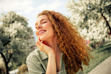 Young red-haired woman enjoying a sunny day outdoors with a beautiful smile in an urban park...