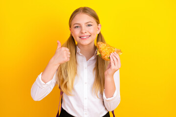 Cheerful schoolgirl enjoys snack with thumbs-up gesture on vibrant yellow backdrop showcasing optimism and positivity