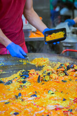 Male vendor making portion of paella at Spanish food festival. Man in gloves serving steaming seafood paella into takeaway container from large skillet at food fair. Concept of paella, street cuisine
