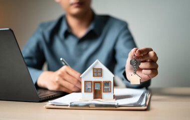 Close-up of hand presenting house-shaped keychain over mortgage papers and model house, symbolizing...