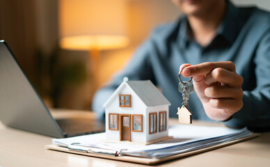Close-up of hand presenting house-shaped keychain over mortgage papers and model house, symbolizing...