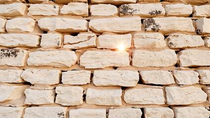 Obraz premium A stone wall made of irregular beige stones, with a small bright light (like sunlight) shining through a gap between two stones.
