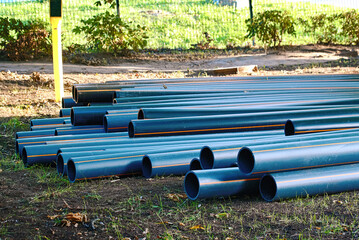 Black hdpe plastic pipes stacked outdoors on the ground in grassy park area prepared for underground electric cable conduit installation at a utility construction site