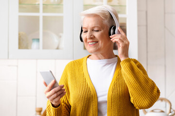 Portrait of happy senior grey-haired woman listening to music on smartphone at home, using wireless...