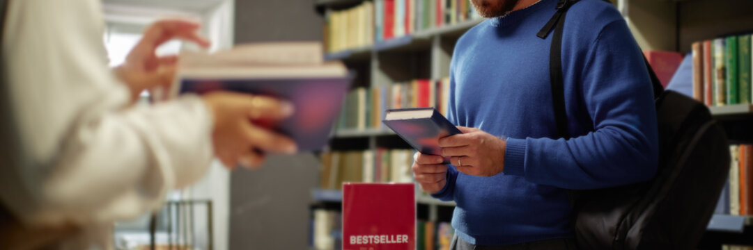 Header of Caucasian young adult man holding book while standing in bookstore near bestseller display, another with feminine hands browsing book in foreground, shelves filled with books in background