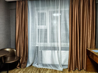 Sheer white curtains gently cover a window in a cozy room with wooden flooring and warm-colored...