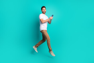 Cheerful young man holding a smartphone while walking confidently against a vibrant teal...