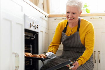 Joyful senior lady in apron opening oven, checking her delicious croissants, cozy kitchen interior,...