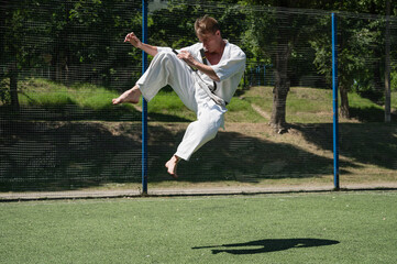 Martial artist performing flying kick outdoors in sunlight.