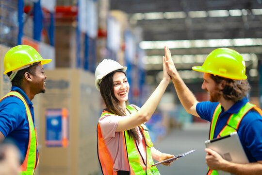 Warehouse workers in safety gear celebrating success with a high five. Smiling colleagues show teamwork, motivation, and achievement in a modern industrial work environment.