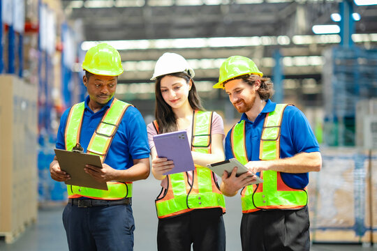 Team of warehouse workers in safety vests and hard hats discussing logistics using digital tablet and clipboard. Industrial teamwork and inventory management in distribution center.
