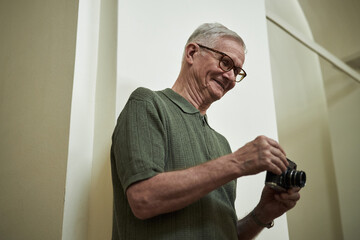 Elderly man examining camera device while standing indoors in relaxed posture, showcasing his...