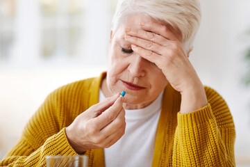 Upset senior woman suffering from headache, touching her temples and taking pill, kitchen interior,...