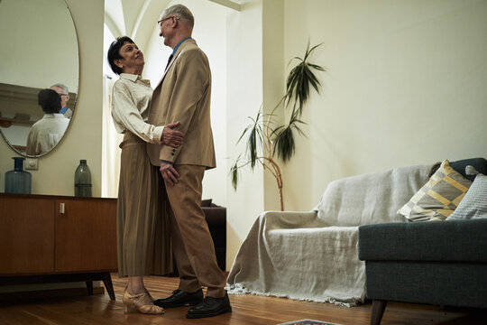 Elderly couple dancing and embracing in modern living room with contemporary furniture and decorations around. Large mirror and potted plant visible in background