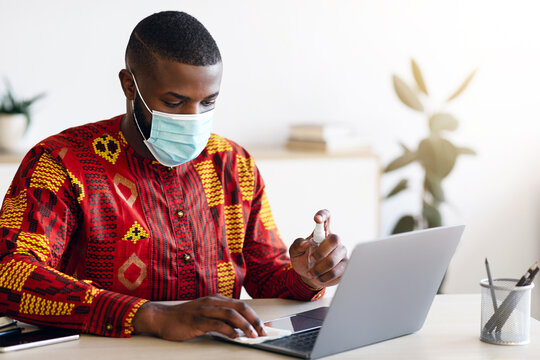 Hygiene On Workplace. African Man In Medical Mask Applying Disinfectant Spray On Laptop Keyboard For Coronavirus Prevention, Black Man In Traditional Clothes Sanitizing Computer Surface, Copy Space - Powered by Adobe