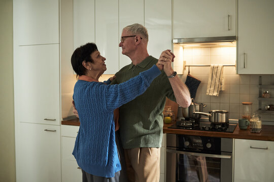 Elderly couple dancing in their modern kitchen, sharing a joyful moment, illuminated by overhead lighting and surrounded by contemporary kitchen appliances, at home
