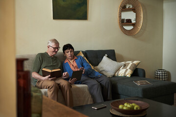 Elderly couple sitting on couch reading books and using tablet in living room creating a serene and...