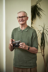 Elderly man with glasses smiling while holding vintage camera, standing indoors in front of potted...