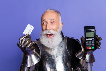 Elderly knight in armor holding a credit card and a POS terminal against a purple background...