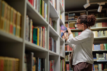Young adult Black woman browsing bookshelves in library, selecting book from shelf, standing in...