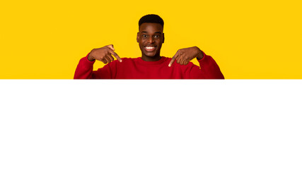 Cheerful handsome black guy pointing down with two fingers, demonstrating free copy space for your design or offer, positive african american man standing isolated over yellow background © Prostock-studio