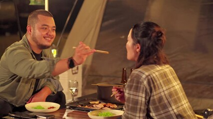 An Asian couple is enjoying a cozy dinner in a camping tent by lantern light. They cook together in front of their tent surrounded by nature near a quiet lake. - Powered by Adobe