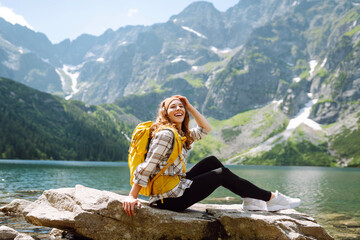 A female hiker with a colorful backpack enjoys the view of a mountain lake and feels freedom. Young...